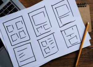 A sheet of paper showing hand-drawn website layout wireframes on a desk beside a laptop and pencil.